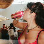 A woman in a bright pink dress drinks from a Strawberry Sunshine can at an outdoor event under a brown mesh canopy.
