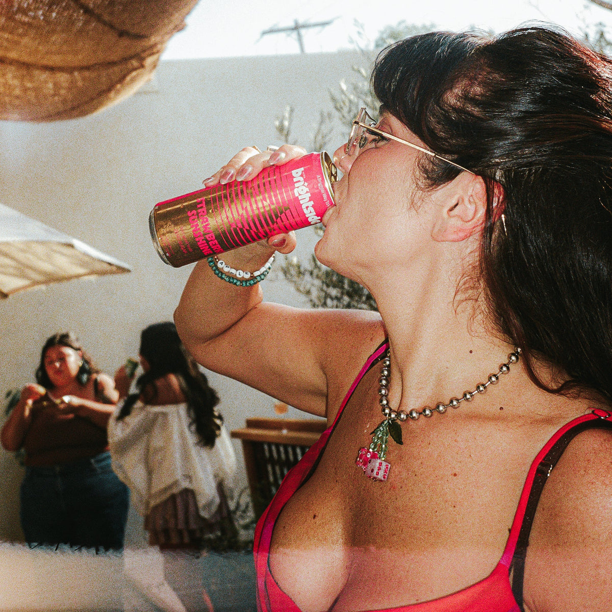 A woman in a bright pink dress drinks from a Strawberry Sunshine can at an outdoor event under a brown mesh canopy.