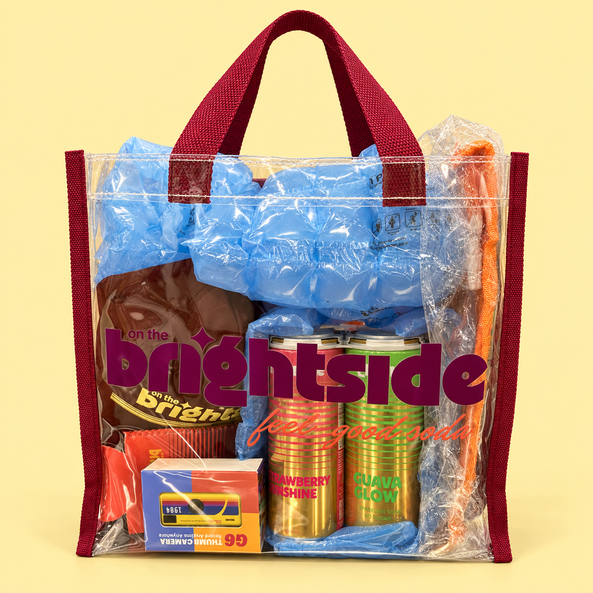 A Brightside clear plastic tote bag with maroon handles and trim, filled with assorted merch including blue packing material, a cap, 2 brightside sodas, a small camera, and a koozie, set against a pale yellow background. The bag has a large 'on the brightside' logo printed on the front.