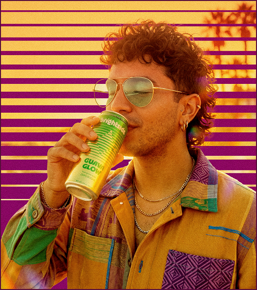 Man in retro clothing drinking can of On The Brightside Guava Glow with palm trees and purple striped background.