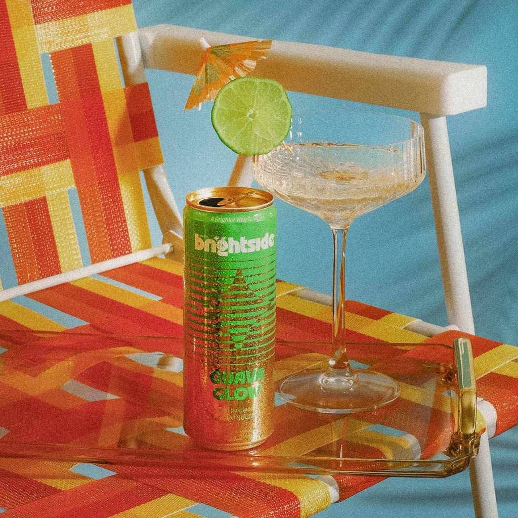 A can of Brightside Guava glow sits on a clear glass tray next to a margarita glass with lime garnish on an outdoor chair with orange, yellow, and red woven fabric.