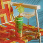 A can of Brightside Guava glow sits on a clear glass tray next to a margarita glass with lime garnish on an outdoor chair with orange, yellow, and red woven fabric.
