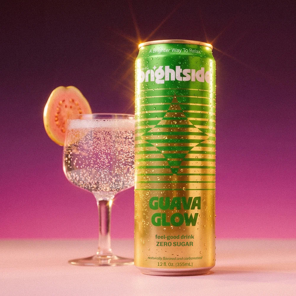 A can of Brightside Guava Glow zero sugar soda stands on a pink surface against a purple-to-pink gradient background, next to a glass of the drink with a guava slice garnish.