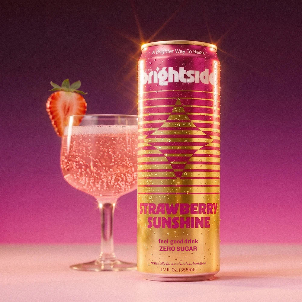 A can of Brightside Strawberry Sunshine zero sugar soda stands next to a stemmed glass filled with it and garnished with a strawberry slice, set against a purple-to-pink gradient background.