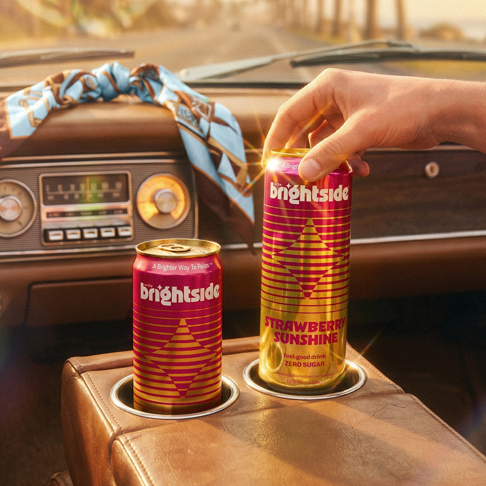 Two Brightside Strawberry Sunshine cans, one being held by a hand and one resting in a cup holder, set in a retro brown leather car interior.