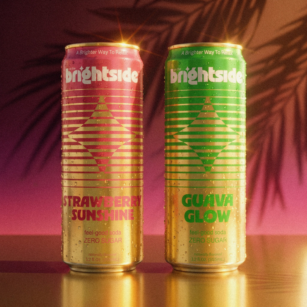 Two On the Brightside cans standing upright on a reflective surface against a warm pink and purple background with palm leaf shadows. The left can is pink and gold with 'Strawberry Sunshine' text, and the right can is green and gold with 'Guava Glow' text.