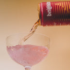 Strawberry Sunshine On the Brightside soft drink being poured into a clear glass, creating a pinkish liquid splash against a warm beige background.